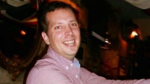 A man stood side on but looking at the camera. He is smiling and holding a tamborine with his arms out stretched. He appears to be in a dark venue with dim lighting.