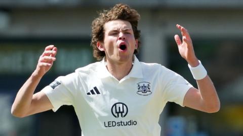 Henry Brookes, wearing a white Adidas cricket shirt with the Gloucestershire county logo, opens his mouth and puts his hands up by the side of his head after bowling a ball in the County Championship Division Two game against Lancashire