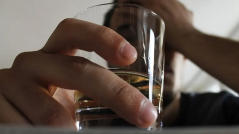 A man holds a drink in his hand close to the camera with his other hand on his head