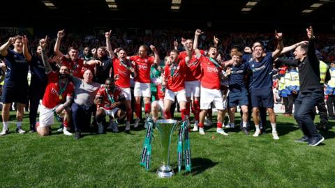 A group of footballers in red and white jumping in the air with hands in the air. Some of the group are wearing navy tops and shorts. They are on a football pitch and there is a silver trophy with blue ribbons on it in front of them.