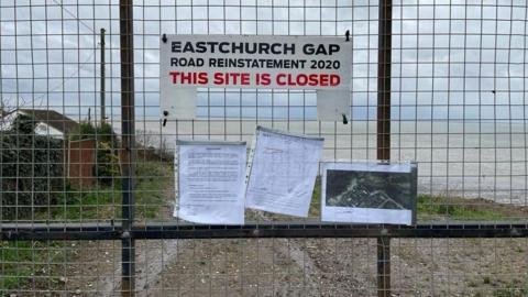 A metal mesh fence with closed sign at Eastchurch Gap with sea and shingle beach in the background