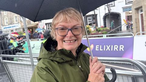 Joan Fletcher, who has short wavy blonde hair and dark-framed glasses and is wearing an olive green waterproof coat with hood, stands smiling in Clitheroe town centre holding a black umbrella