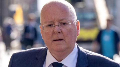Close-up of Peter Murrell, a bald man wearing glasses, a dark suit, white shirt, and tie, walking outdoors on a sunlit city street with a blurred background of pedestrians and buildings.