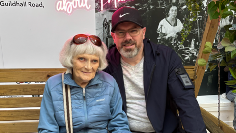 A woman in a blue puffer jacket and jeans sits on a wooden bench. Next to her is a man in a navy coat, a white jumper and grey jeans. They are looking at the camera and on the left hand side are lots of flowers. 