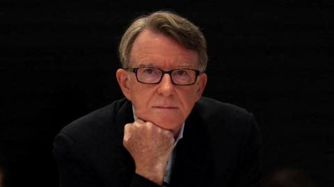 Peter Mandelson is resting his chin on his fist. He has short brown hair and black square-framed glasses.