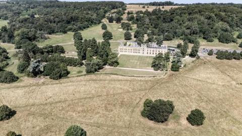 A drone view of Ashton Court estate with large areas of grassland showing light brown instead of green. Small trees are dotted around the landscape and the lines of footpaths through the landscape are also visible
