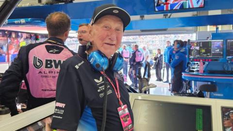 An older man wearing a black cap, blue headphones, and a black shirt stands in a motorsport pit area. Behind him, team members in pink and black uniforms work near monitors, with spectators visible through the open garage.