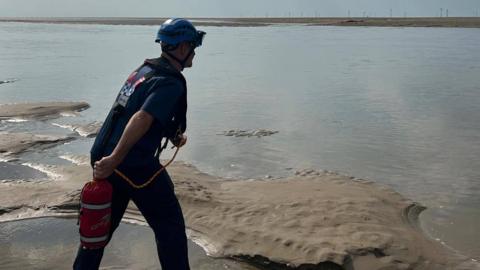 A rescue officer in blue uniform and helmet appears to throw out a safety line from the sands acorss the water at Leasowe.