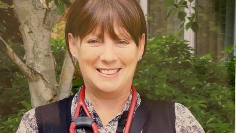 Beverley Pedley, stood in what appears to be a garden, with a stethoscope around her neck and a blue gilet and floral blouse.