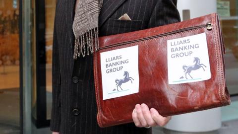 A man holds up a briefcase saying "Liars Banking Group" on it