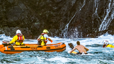The picture shows a sea rescue operation involving the RNLI. Three rescuers are in action - one in the water wearing yellow and red RNLI life jackets, and two in an orange inflatable rescue boat marked Y242 along with a paddleboarder.