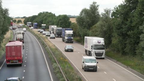 A view of a dual carriageway with a central barrier separating traffic moving in opposite directions. Dense green trees and bushes line both sides of the road, and the sky above is overcast with grey clouds.