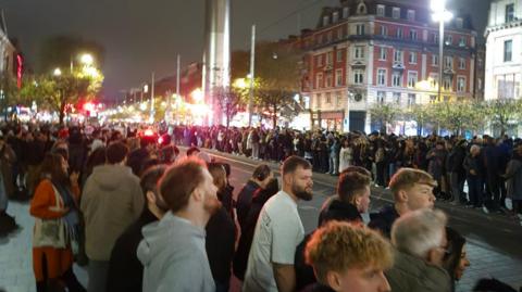 Crowds lining the streets in Dublin City Centre, with the Dublin Spire in the background. It is night time, and the streets are lit.