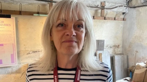 A woman with long grey hair, a blue and white striped top and a red lanyard around her neck is standing in a room in front of a stone wall with concrete-type slabs on the floor.