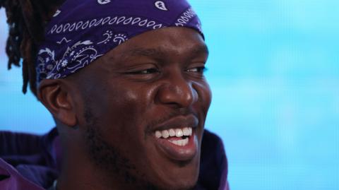 KSI, wearing a blue and white bandana, laughing at a boxing press conference  last year