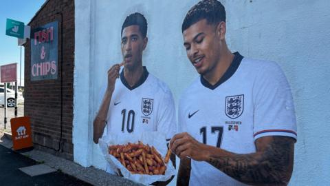 A mural on the side of a white building shows England footballers Jude Bellingham and Morgan Rogers eating from a bowl of orange chips.