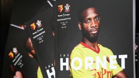 A scattered pile of football programmes featuring a player in a yellow and red kit with arms crossed on the cover.