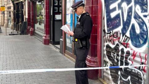 A police officer stands behind police tape on a city street in central London, reading notes outside a closed shop with graffiti on its shutter, while nearby streets are quiet and cordoned off.