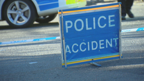 A blue police accident sign in front of a line of police tape and the wheel of a police car.