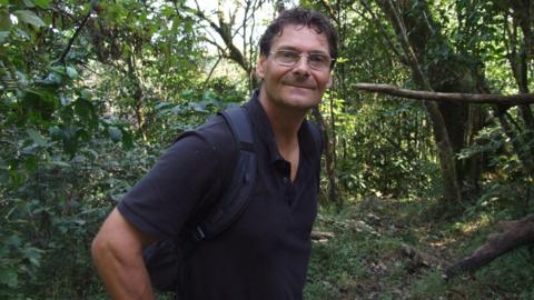 A man with short curly dark hair and silver-rimmed glasses smiles at the camera. He wears a black polo shirt and a blue rucksack. Behind him, a dense green forest vegetation can be seen.