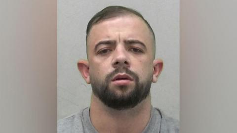 Mugshot of a young man with closely cropped dark hair and a dark beard. He is wearing a grey t-shirt or sweatshirt, and has a serious expression.