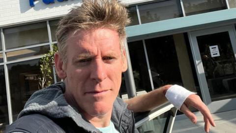 A man with short grey hair is resting his bandaged hand while standing in front of a hospital building.