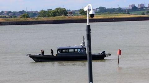 A black boat on a river. There is someone on the front looking through binoculars.