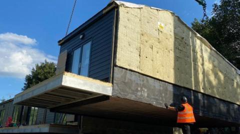 One of the six properties being assembled in a row next to the others. They are built off-site. A crane lifts a near-complete house to its required location whilst a man in an orange high-visibility jacket supervises.