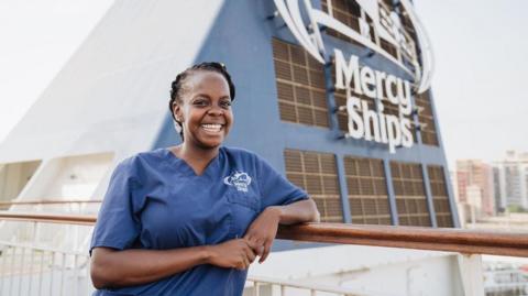 A woman in a blue Mercy Ships uniform stands on the deck of a hospital ship, leaning on a railing, with the charity logo visible behind.