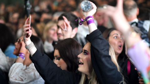 A young female clubber points her phone towards the stage, surrounded by other clubbers