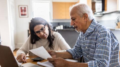 Older man and woman sit at table looking intently at a piece of paper the man is holding. On the table in front of them is an open laptop, a pad of paper, numerous sheets of loose paper, and a calculator