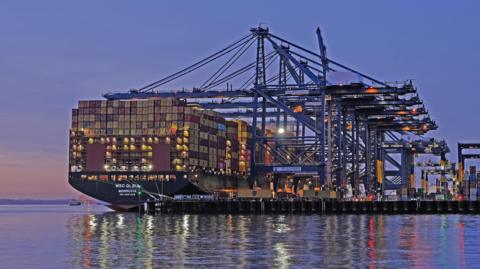 A large container ship docked at the Port of Felixstowe at sunset. The lights from the ship and dock are reflected in the water.