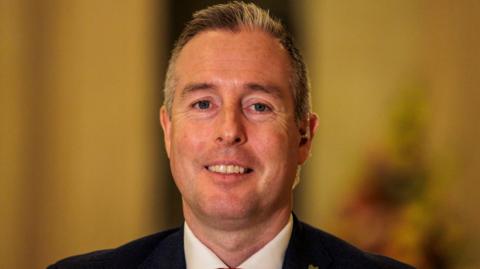 Paul Givan smiling at the camera, he has greyish black hair, he is wearing a black blazer and white shirt
