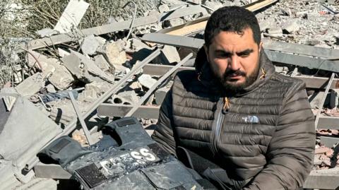 A man holding a damaged flak jacket with "press" written on it, rubble is visible in the background