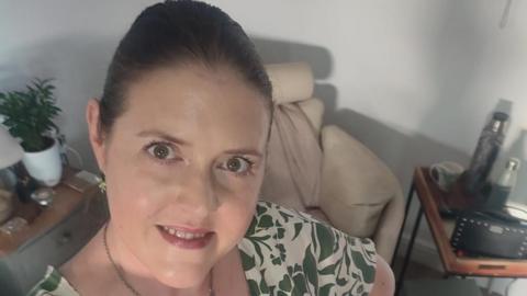 Rachael Johnson who is 41 years old. She is smiling and looking up at the camera. She has hazel/brown eyes and is wearing a short sleeve summer dress which is cream with a khaki leave pattern.