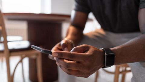 A man sits in a kitchen typing on his smartphone.