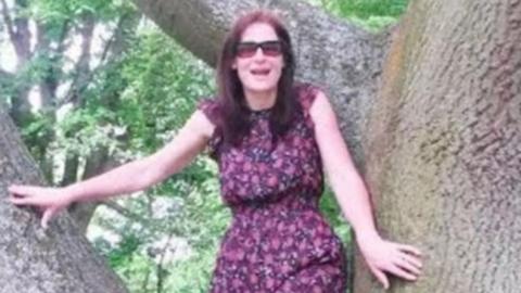 A middle-aged woman with long reddish hair and wearing a floral dress and sunglasses stands smiling between two big tree branches. 