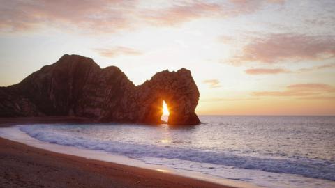 Durdle Door is a huge, natural limestone arch on the Dorset coast. The sun can be seen shining through the archway casting a yellow glow on the sand.