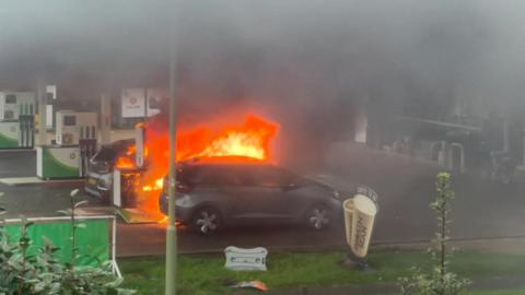 Two cars at a BP petrol station with a fire blazing on the pumps between them