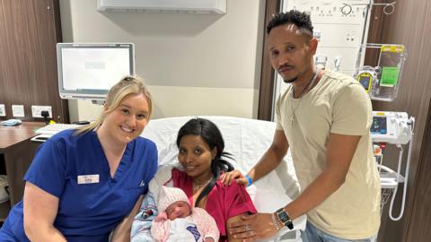 A midwife in a blue uniform is smiling, sat on the side a hospital bed with a woman in a pink outfit who is also smiling whilst holding a newborn baby. Stood alongside the woman is a man in a beige t-shirt who has his hands placed on her arm and shoulder. There is hospital equipment in the background.