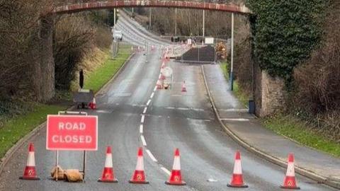 Kewaigue Hill with the railway bridge running over it. There are traffic cones across the carriageway and a red road closed sign, with the work site in the background.