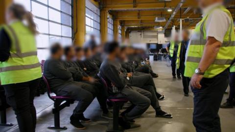Staff in dark uniforms and hi-vis jackets look on as two rows of people in track suits and sandals sit waiting on two long rows of chairs in a large room that looks like a warehouse.     