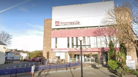 Thameside Theatre seen from the front. It is a brown brick building with large white and faded red blocks on the front, with the word thameside all in lower case. is is next to a road and pedestrianised area with railings and an old-fashioned lamp post. Some trees and shrubs are placed to the right of the theatre.