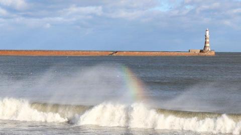 A small rainbow forms in a breaking wave with flat grey sea behind, a lighthouse and blue cloudy skies