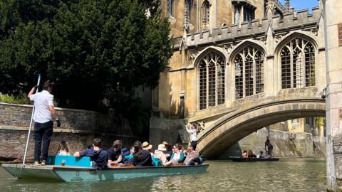 A flat-bottomed punt glides along the River Cam beneath an ornate stone bridge with Gothic-style arches. The boat is steered by a standing guide using a long pole, while several passengers sit and enjoy the view. Another punt is visible in the background, and the scene is framed by the historic Cambridge University buildings.