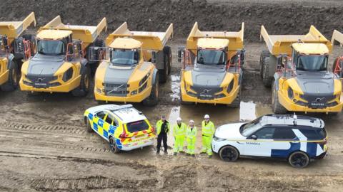 An aerial view of five large yellow vehicles. In front are four people in high vis. Next to them is a police car. 