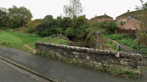 A wall is fencing off a stream next to a road with houses in the background.