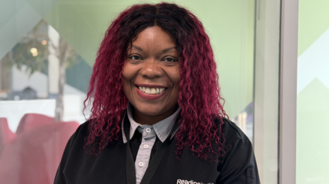 Alma Cox is smiling as she stands in the BBC offices, wearing her Reading Buses shirt, with his black and has a grey collar.