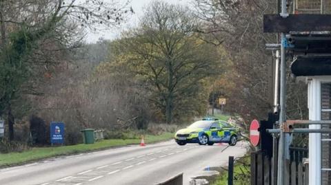A road surrounded by trees with a police car parked on the road cordoning it off.