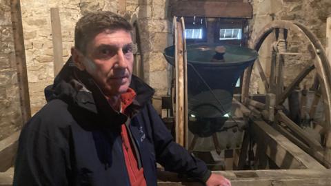 A man stands inside what appears to be an old stone bell tower. The walls around him are built from large, weathered blocks of stone, giving the space a distinctly medieval or early‑historic feel.